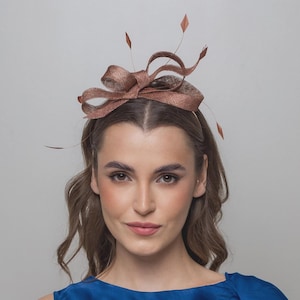 May include: A copper-coloured fascinator headpiece featuring a structured bow design made from a woven, textured fabric. The accessory is accented with thin, delicate wire stems topped with small, diamond-shaped copper ornaments extending outward.