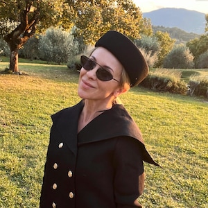 Pode incluir: Uma mulher usa um chapéu preto e óculos de sol com um casaco preto com botões dourados. O casaco tem uma gola única. O fundo mostra um campo gramado verde e árvores.