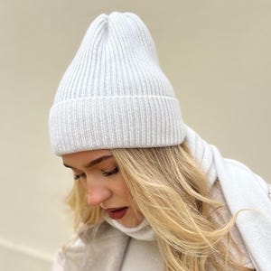 May include: A white knitted beanie with a ribbed texture. The beanie is worn by a person with long blonde hair.
