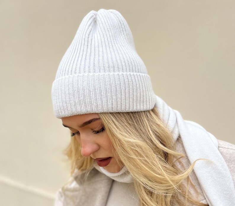 May include: A white knitted beanie with a ribbed texture. The beanie is worn by a person with long blonde hair.