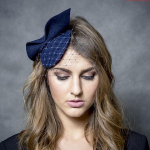 Pode incluir: Uma mulher usando um chapéu de feltro azul marinho com véu. O chapéu é decorado com um laço e é usado do lado da cabeça.