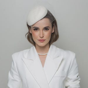Può includere: Un cappello pillbox bianco caratterizzato da un delicato velo a rete fine che copre la parte superiore del viso. Il cappello è abbinato a un blazer bianco e a una classica collana di perle, creando un insieme coordinato dall'estetica monocromatica.