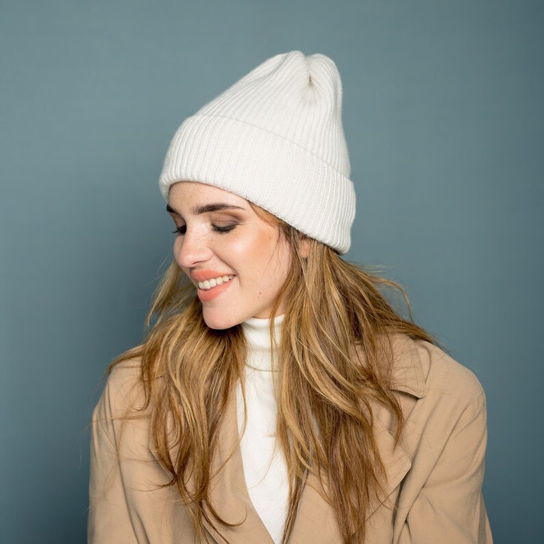 May include: A woman with long blonde hair is wearing a white knit beanie and a beige coat. She is looking down and smiling.