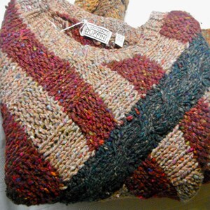 Könnte beinhalten: Ein mehrfarbiger Strickpullover mit einem diagonalen Muster in Braun-, Beige-, Rot- und Blautönen. Der Pullover hat einen Rundhalsausschnitt und ist aus einem dicken, strukturierten Garn gefertigt. Das Etikett lautet "Anfrano Ruffini".