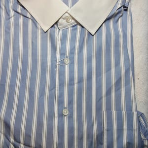 Pode incluir: Uma camisa de vestir azul e branca listrada com colarinho branco. A camisa tem uma etiqueta que diz "Saks Fifth Avenue".