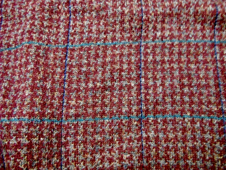 May include: Close-up of a red, brown, and blue plaid fabric with a houndstooth pattern. The fabric has a textured, woven appearance.