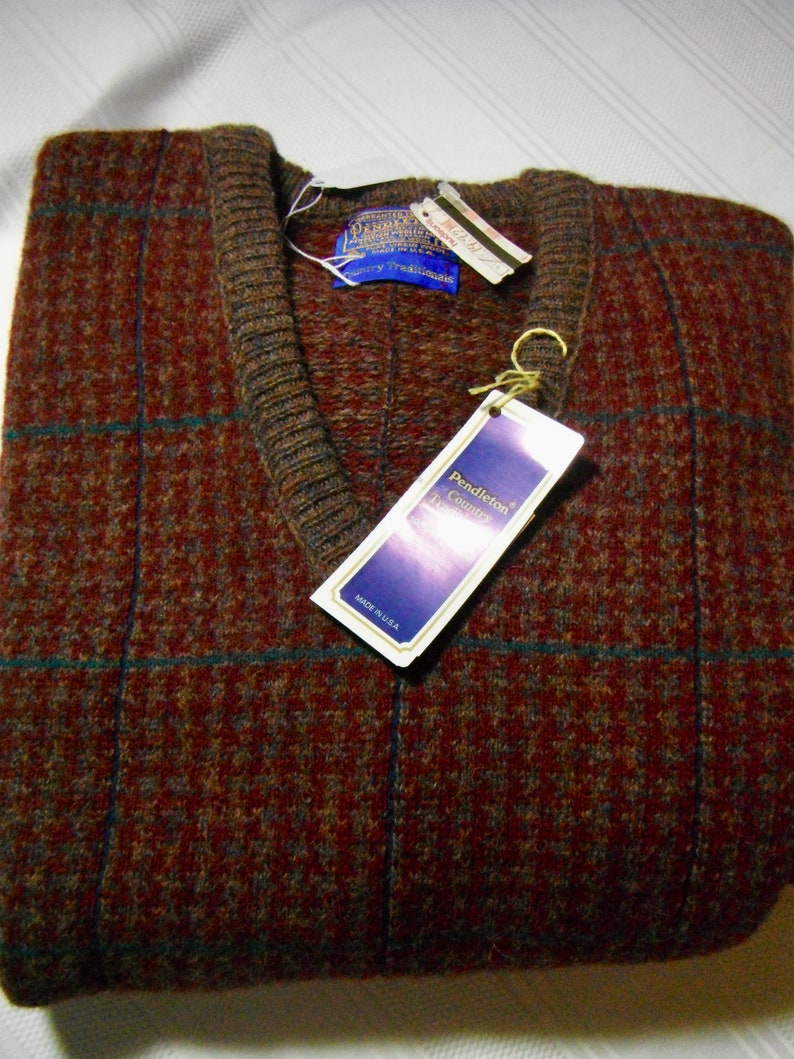 May include: A brown and blue plaid wool sweater vest with a V-neck. The sweater has a Pendleton Country Traditions label attached to it. The label is blue with white text and says "Made in U.S.A."