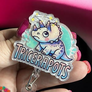 May include: A sparkly blue and white badge holder with a cartoon illustration of a blue and purple triceratops with a dazed expression. The text "TRICERAPOTS" is written in a glittery white font.