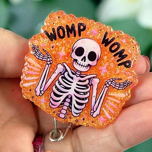 May include: Orange glitter badge holder with a skeleton wearing a shirt that says "Womp Womp".