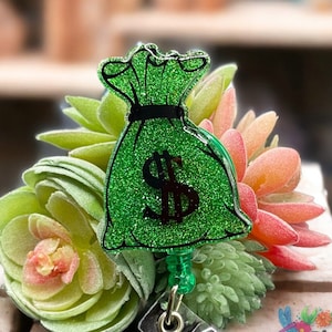 May include: Green glitter money bag charm with a black dollar sign on a retractable badge reel. The reel is green and has a green beaded end cap.