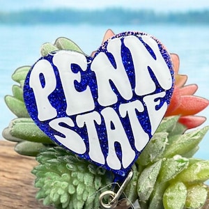 May include: A heart-shaped badge reel with the words "PENN STATE" in white lettering on a blue glitter background. The reel is attached to a clear plastic clip with a blue gem. The badge is surrounded by green and red succulents.