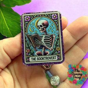 May include: A purple glitter badge holder with a skeleton reading a book. The image is surrounded by a floral border and the text "The Booktrovert" is at the bottom.