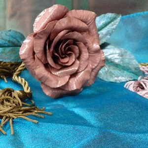 May include: A single, large, pink rose made of metal with green leaves. The rose is lying on a blue fabric surface.