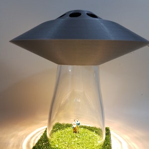 May include: A unique lamp featuring a gray UFO-shaped top above a clear glass enclosure. Inside, a miniature couple stands on a patch of green grass. The lamp base is illuminated, creating a warm glow.
