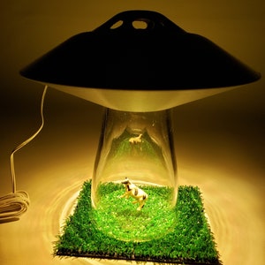 May include: A unique lamp with a black saucer-shaped top and a clear glass base. Inside the glass, a miniature cow figurine stands on a patch of green artificial grass. The lamp's design resembles a UFO.