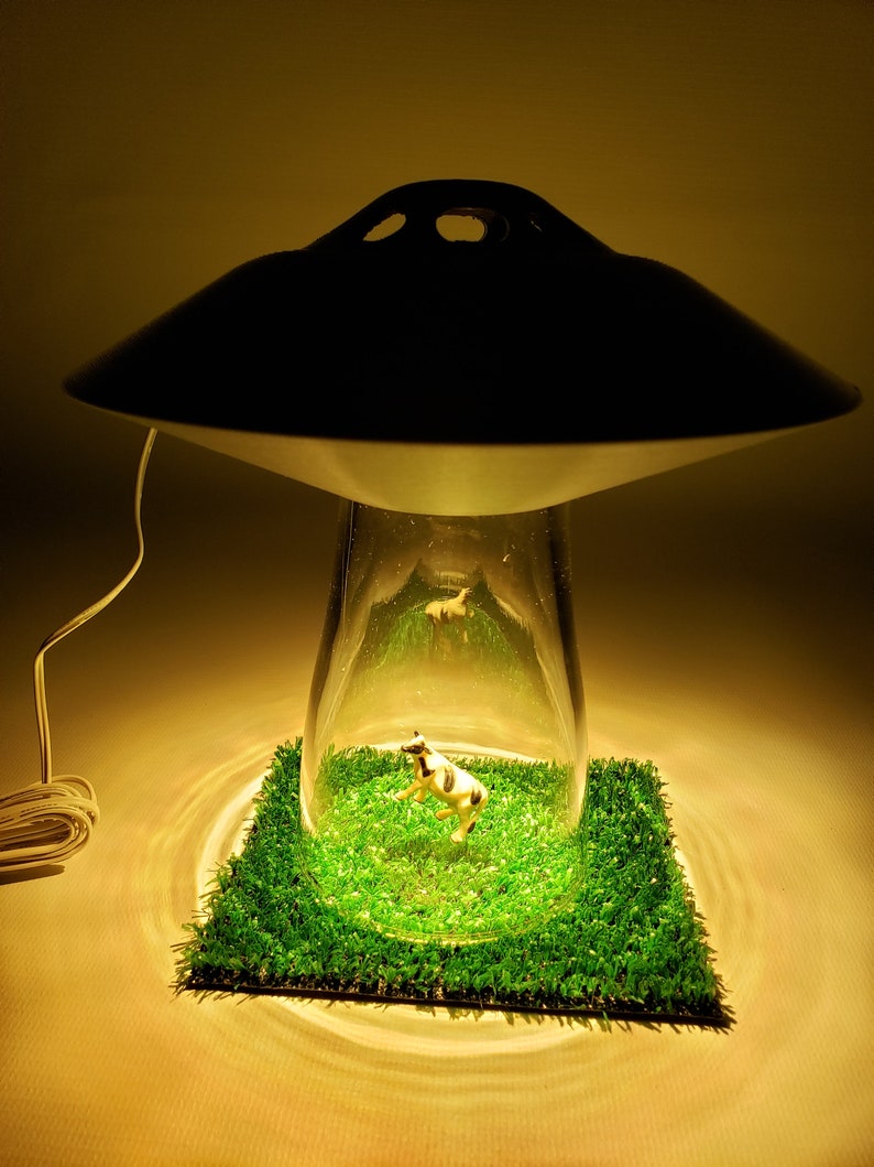 May include: A unique lamp with a black saucer-shaped top and a clear glass base. Inside the glass, a miniature cow figurine stands on a patch of green artificial grass. The lamp's design resembles a UFO.