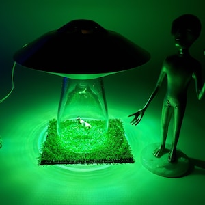May include: A novelty lamp featuring a black UFO design with a clear glass base. Inside, a miniature cow figurine rests on artificial green grass. A green alien figure stands nearby, illuminated by green light.