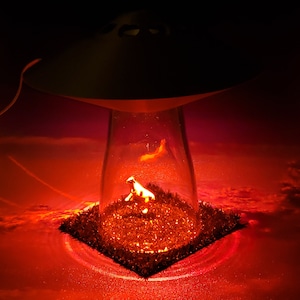 May include: A novelty lamp featuring a UFO design with a clear glass enclosure. Inside, a miniature cow figurine stands on a patch of artificial grass, illuminated by a red glow. The background suggests a sunset.
