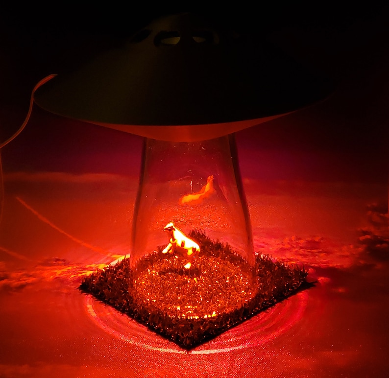May include: A novelty lamp featuring a UFO design with a clear glass enclosure. Inside, a miniature cow figurine stands on a patch of artificial grass, illuminated by a red glow. The background suggests a sunset.