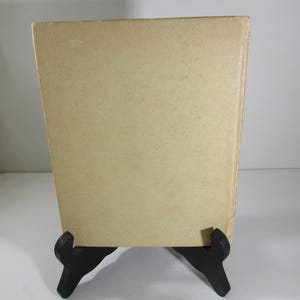 May include: The back cover of a vintage paperback book with a plain, off-white cover. The book is on a black book stand.