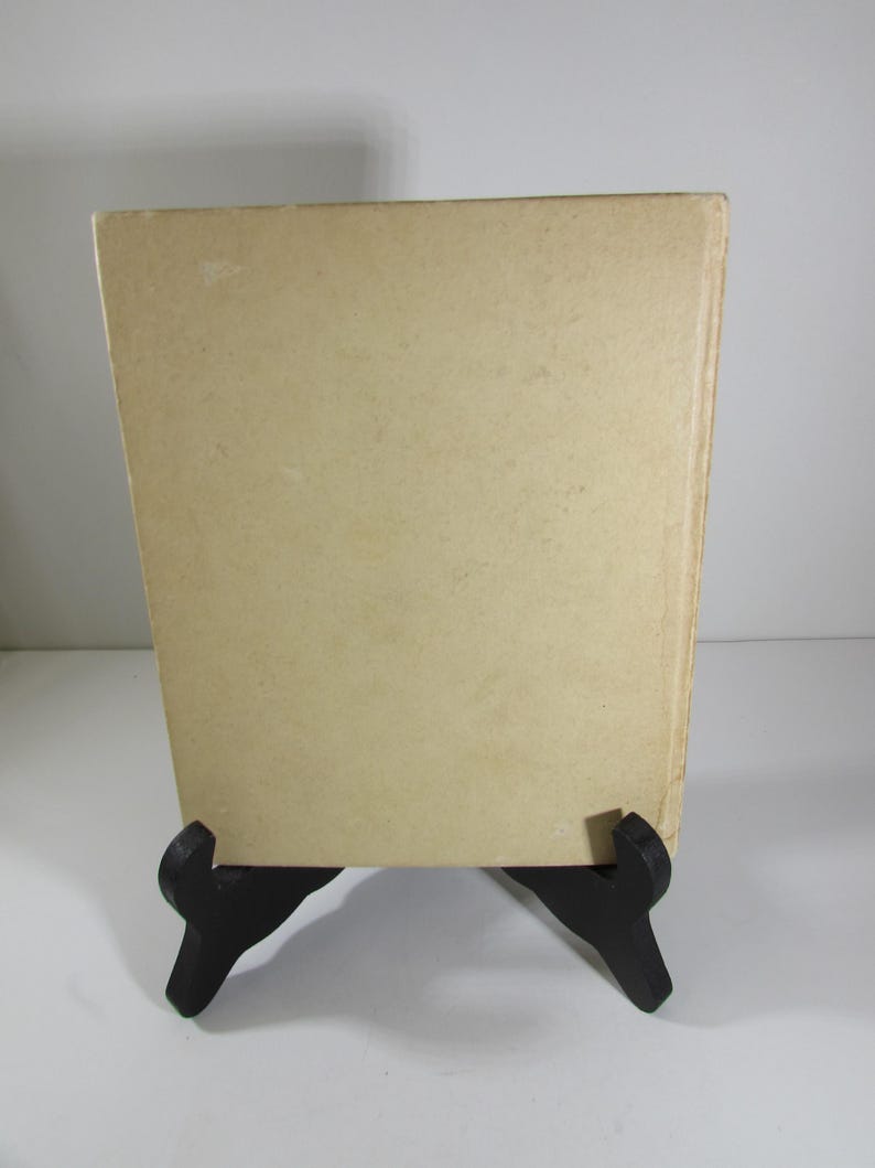 May include: The back cover of a vintage paperback book with a plain, off-white cover. The book is on a black book stand.