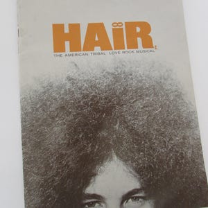 May include: A rectangular program booklet for the musical HAIR. The cover features the orange text HAIR THE AMERICAN TRIBAL LOVE ROCK MUSICAL above a black and white photograph of a person with voluminous curly hair and visible eyes.
