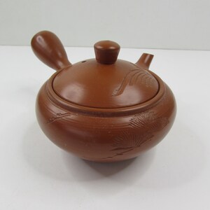 May include: A small, brown clay teapot with a lid and a handle. The teapot has a floral design carved into the side.