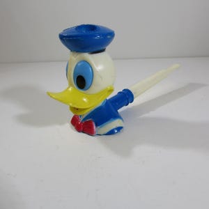 May include: A vintage Donald Duck figurine. The figure is primarily white, with a yellow beak, blue eyes, and a blue sailor hat. It wears a blue sailor shirt and a red bow tie. The figurine has a white, pen-like extension.