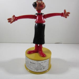 May include: A vintage Olive Oyl push button puppet toy. The figure has a red top and arms, a black skirt, and red shoes. The base is yellow with text that reads "Olive Oyl Push Button Puppet".