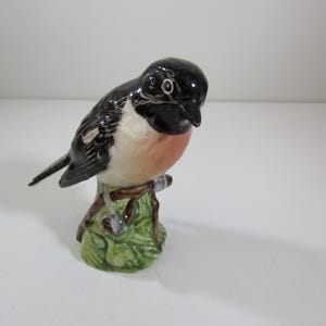 May include: A ceramic bird figurine with a black head and wings, a white chest, and a reddish-orange breast. The bird is perched on a brown branch with green leaves. The figurine is a decorative item.
