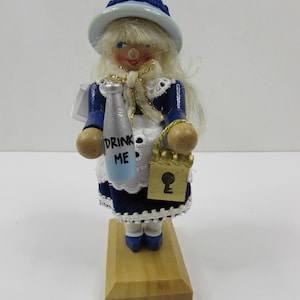May include: A wooden doll dressed in a blue and white outfit, holding a bottle with "DRINK ME" on it and a gold-colored purse with a keyhole. The doll has blonde hair and a blue hat. It stands on a wooden base.