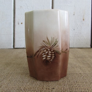 May include: A white ceramic vase with a brown and green pine cone design. The vase has a hexagonal shape and a smooth, glossy finish.