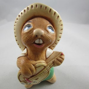 May include: A ceramic figurine of a brown rodent wearing a beige cowboy hat and playing a guitar. The rodent has blue eyes, a pink mouth, and is wearing a white shirt and green shorts. The guitar is beige with red accents.