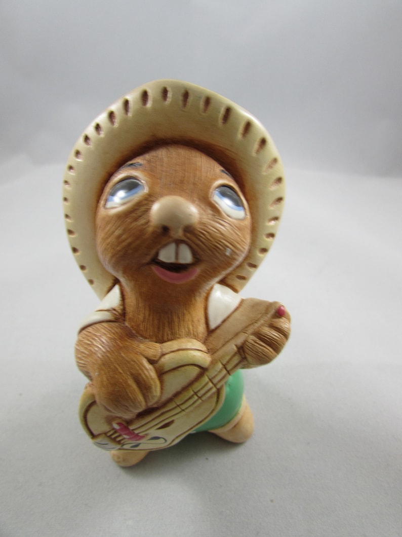 May include: A ceramic figurine of a brown rodent wearing a beige cowboy hat and playing a guitar. The rodent has blue eyes, a pink mouth, and is wearing a white shirt and green shorts. The guitar is beige with red accents.