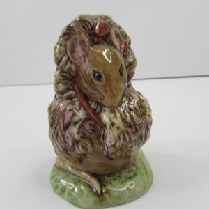 May include: A small ceramic figurine of a brown mouse wearing a pink flower crown and holding a bouquet of pink flowers. The mouse is sitting on a green base.