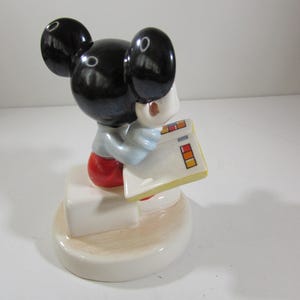 May include: A ceramic figurine of Mickey Mouse, wearing a light blue shirt, red shorts, and black ears. Mickey is holding a white notepad with colorful rectangles. The figurine is on a white base.