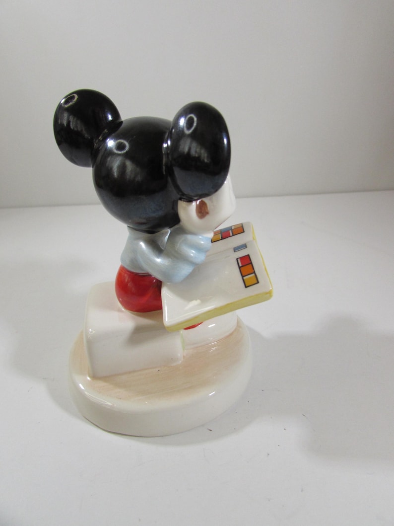 May include: A ceramic figurine of Mickey Mouse, wearing a light blue shirt, red shorts, and black ears. Mickey is holding a white notepad with colorful rectangles. The figurine is on a white base.
