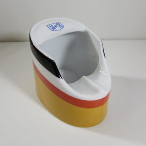 May include: A white ceramic ashtray with a black, white, and red stripe design. The ashtray has a logo that says "HAPAG" in blue.