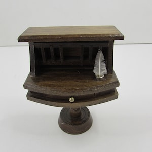 May include: A small wooden desk with a drawer and a hinged top. The desk has a turned pedestal base and a small white feather is resting on the top.