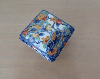 May include: Square ceramic trinket box with a floral design in blue, red, yellow, and gold on a white background.