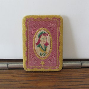 May include: A vintage Girl Scout patch with a gold border and a red background. The patch features a girl scout in a green uniform with a gold pin and a gold four-leaf clover. The text "Girl Scout" is printed in gold letters on the top and bottom of the patch.
