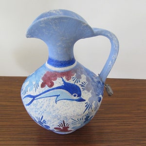 May include: A blue and white ceramic pitcher with a dolphin design. The pitcher has a wide mouth and a handle. The dolphin is painted in blue and white with red accents.