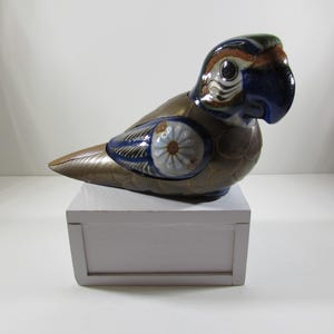 May include: A ceramic parrot figurine with blue, gold, and white painted details. The parrot is perched on a white wooden base.
