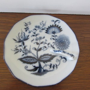 May include: A white porcelain saucer with a blue floral design and a handle. The saucer has a scalloped edge and is decorated with a stylized floral pattern in shades of blue.