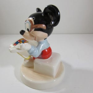 May include: A ceramic figurine of Mickey Mouse wearing goggles and red shorts, seated on a white block. The black-eared mouse is holding a colorful square. The base is a light beige color.