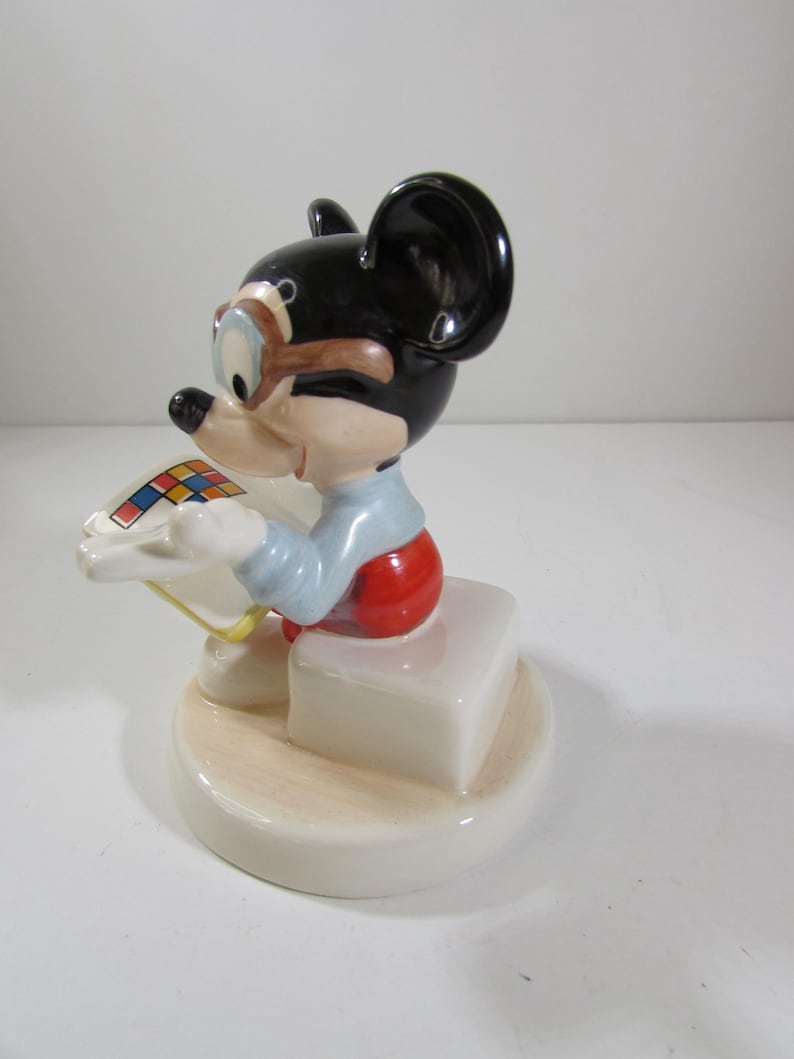 May include: A ceramic figurine of Mickey Mouse wearing goggles and red shorts, seated on a white block. The black-eared mouse is holding a colorful square. The base is a light beige color.