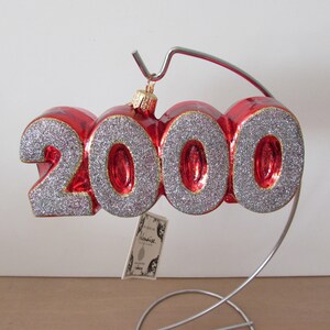May include: Red glass Christmas ornament shaped like the year 2000 with silver glitter. The ornament is on a metal stand.