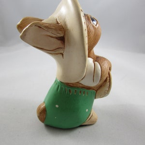 May include: A ceramic figurine of a rabbit wearing a cream-colored hat and green overalls with white polka dots. The rabbit has brown fur, blue eyes, and is holding its hands together. The figurine is a decorative item.