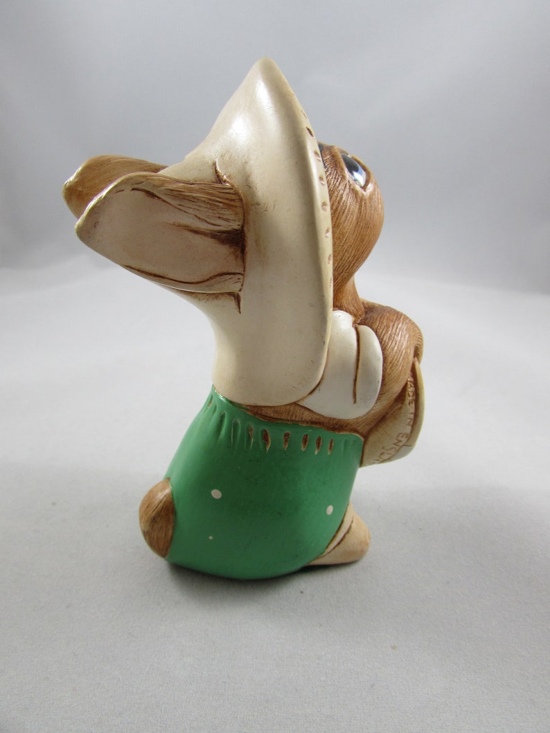 May include: A ceramic figurine of a rabbit wearing a cream-colored hat and green overalls with white polka dots. The rabbit has brown fur, blue eyes, and is holding its hands together. The figurine is a decorative item.