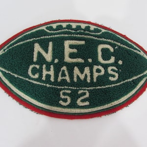 May include: An oval-shaped embroidered patch with a dark green background and a red border. The patch features the white text N.E.C. CHAMPS 52, arranged in a football-like design with white stitching details outlining the shape.